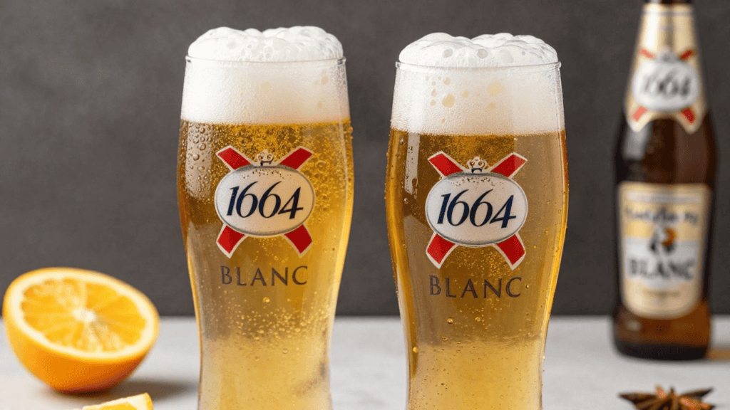 Two frosty glasses of Kronenbourg 1664 Blanc beer with foamy tops, a halved orange on the left, and a bottle in the background.