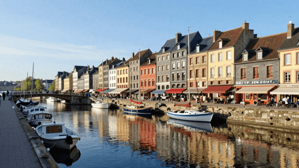 Colorful townhouses line a canal with boats moored along the edge and people strolling the promenade.