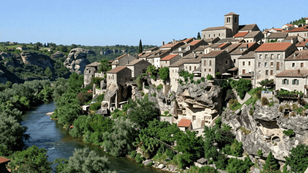 Cliffside village with stone houses and red-tiled roofs perched above a winding river, surrounded by trees under a blue sky.