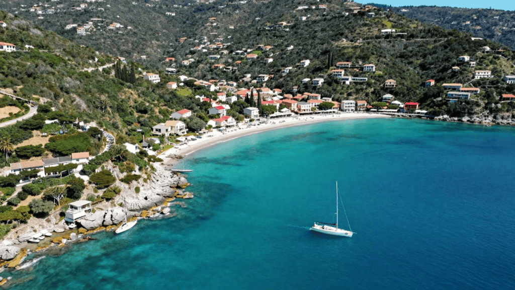 A coastal hillside village with colorful houses along a white-sand beach and a sailboat anchored in clear turquoise bay