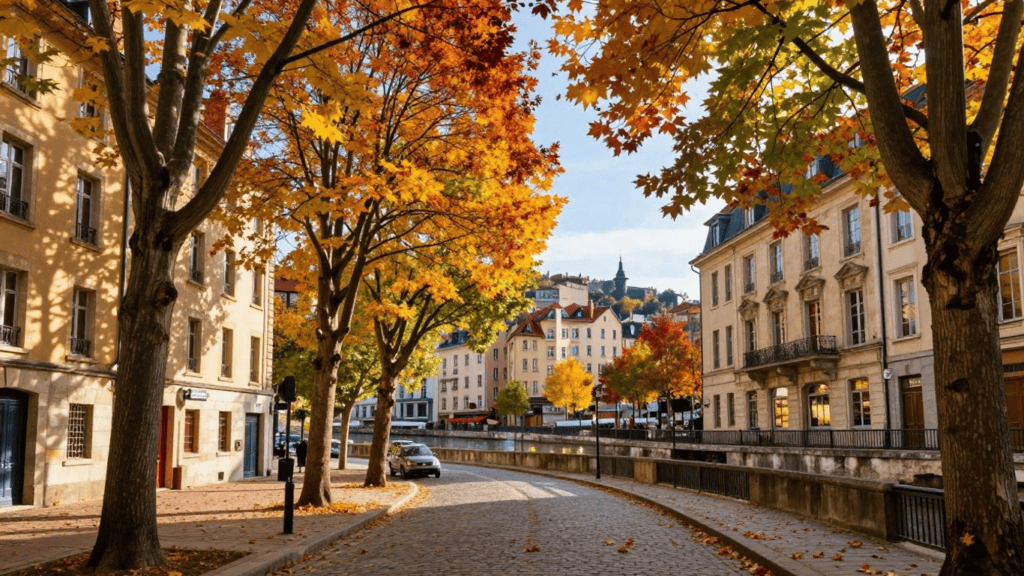 Quaint cobblestone street lined with fall-colored trees and classic European buildings along a canal and railing.]