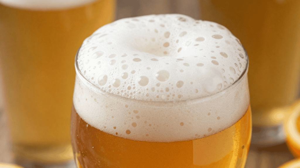 Beer glass with creamy foamy head and amber liquid, surrounded by others on a wooden surface.