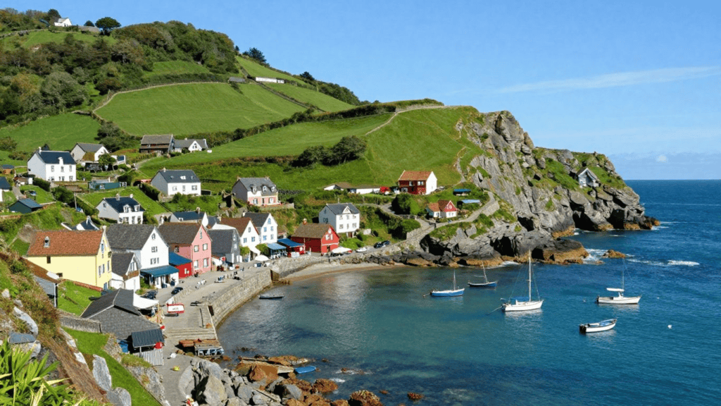Colorful seaside village with white, pink, and red houses on a green hillside beside a calm harbor and rocky coastline.