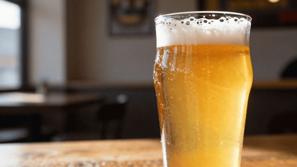 Tall glass of frothy golden beer on a wooden bar counter, close up with bubbles at the top.