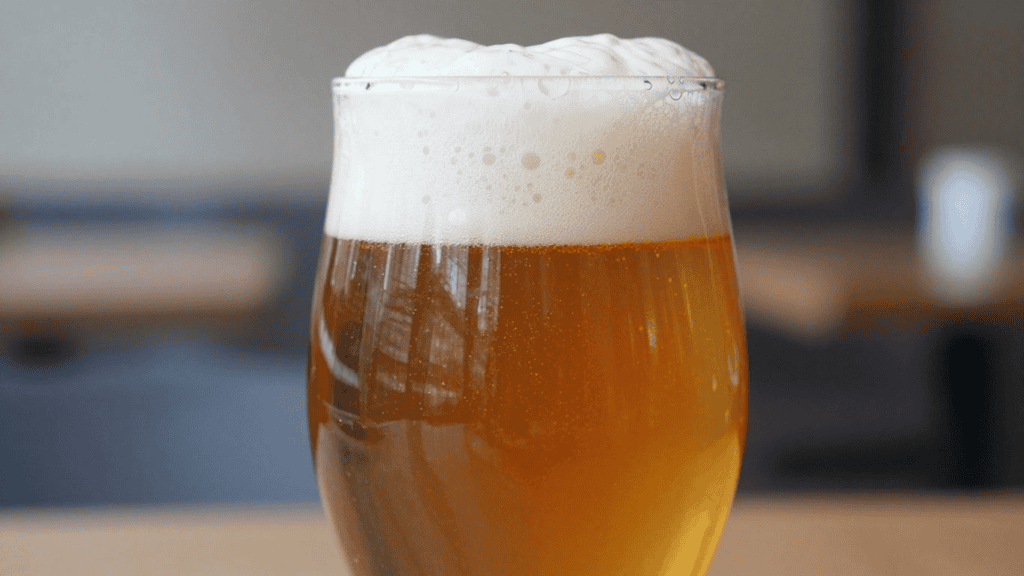 Glass of amber beer with a foamy white head on a blurred background, in a tall tulip glass.