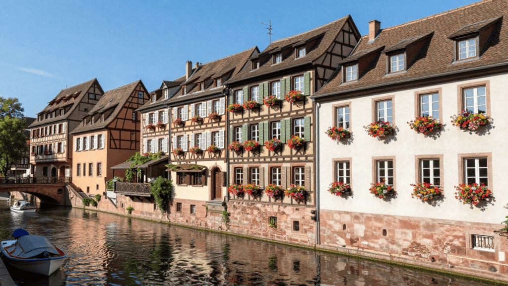 Colorful traditional European houses line a narrow canal with flower-filled window boxes under a clear blue sky.