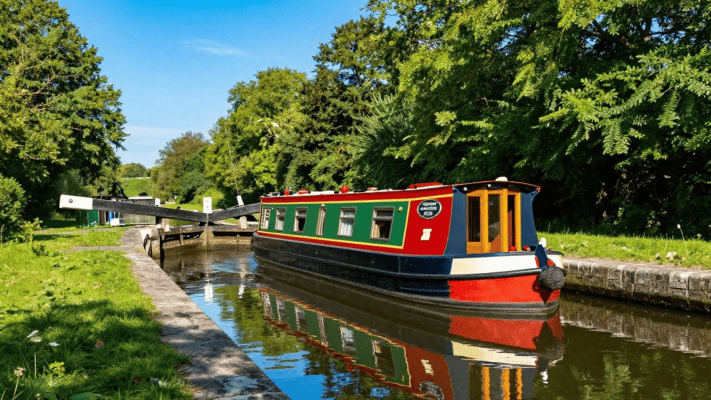 Colorful narrowboat on a calm canal beside a green park, with a lock visible in the distance.