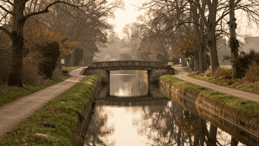 Stone arch bridge over a calm canal, with dirt paths and leafless trees along both banks in autumnal tones.