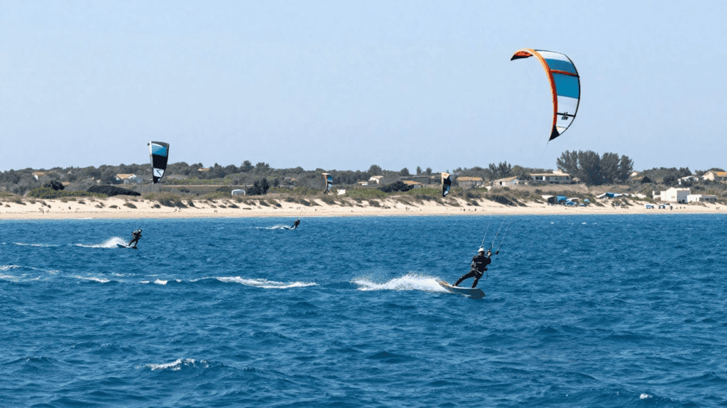 Kitesurfers riding the blue ocean near a sandy coastline with houses in the background and a large colorful kite overhead.