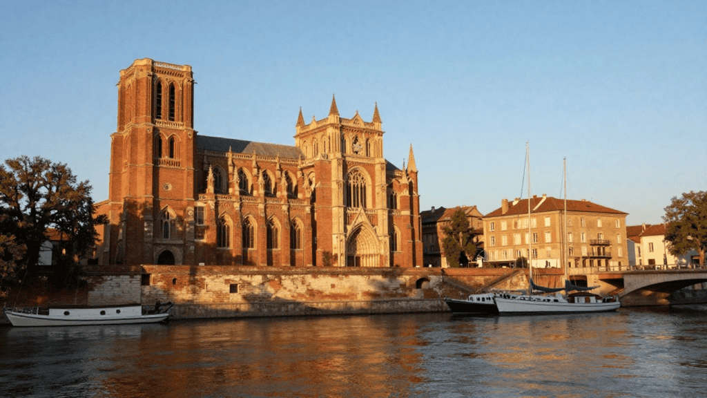 Brick Gothic cathedral by the river with boats docked along the bank at golden hour.