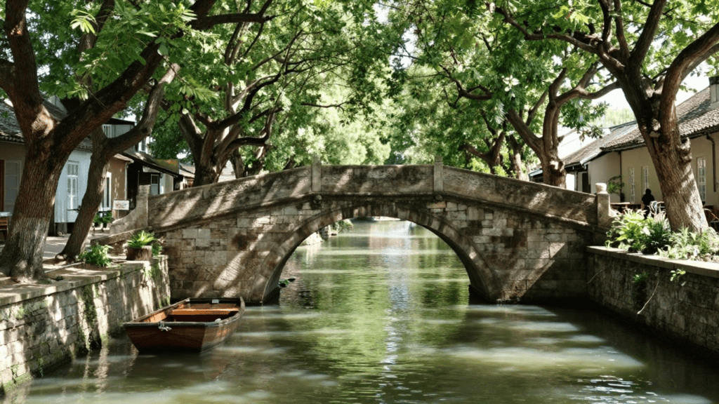 Stone arched bridge over a calm canal framed by leafy trees, with a wooden boat docked along the stone quay on the left.