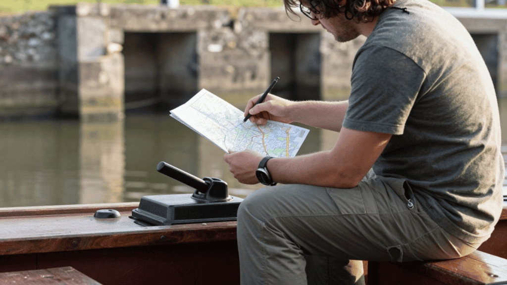 Person sitting in a wooden boat on a calm river, studying a folded map with a pen in hand.