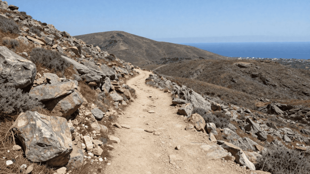 Dusty hiking trail winds along a rocky hillside with sparse shrubs, mountains in the distance and the sea on the horizon