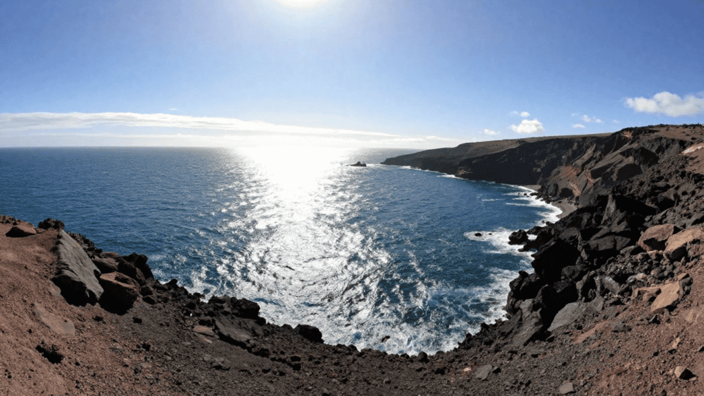 Cliffside coastline with jagged volcanic rocks, blue ocean, and sunlit water sparkle on the horizon.