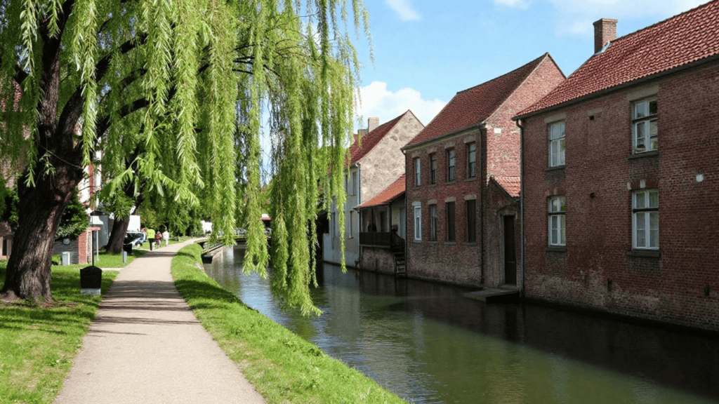 Path beside a narrow canal with brick houses on the right and a large weeping willow arching over the walkway.