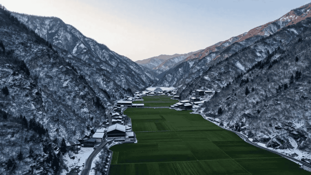 Winter aerial view of a snow-covered alpine valley with green fields and a cluster of chalet-style houses along a river/road between the mountains.