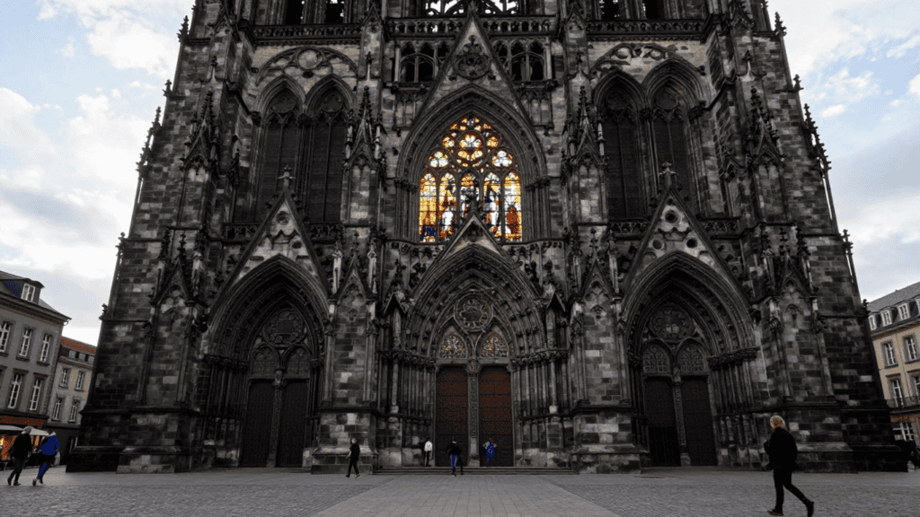 Front of a dark Gothic cathedral with three arched doors, intricate stone carvings, and a colorful stained-glass window above.