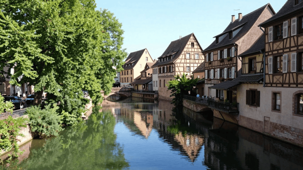 Canal lined with timber-framed houses and a stone bridge, reflected in calm water.”,