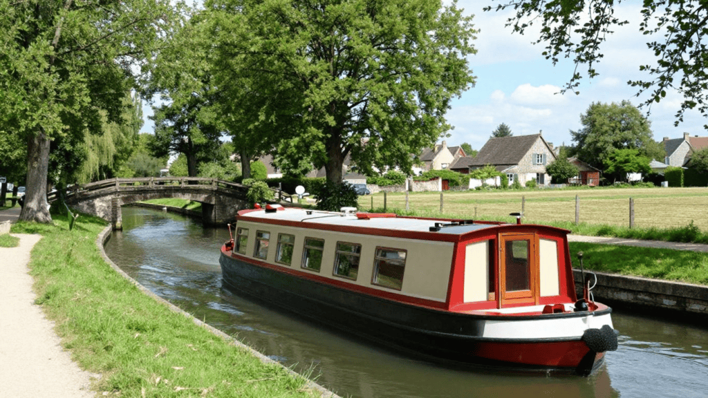 Red and cream canal boat sailing along a narrow canal, with a stone arched bridge, green banks, and a village of thatched-roof houses in the background.