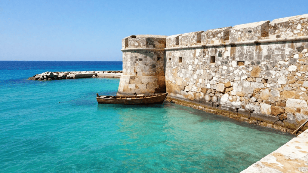 Stone fortress wall along a calm turquoise harbor, with a small wooden boat moored beside it and clear blue sea beyond.