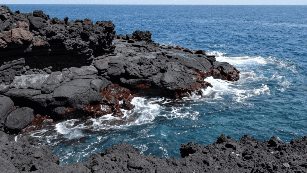 Jagged black lava rock shoreline meeting the blue ocean as waves crash and foam along the coast, under a clear sky.