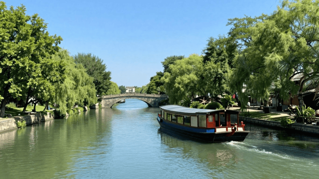 A boat gliding along a narrow canal under a stone arch bridge, with lush trees lining both banks on a sunny day.