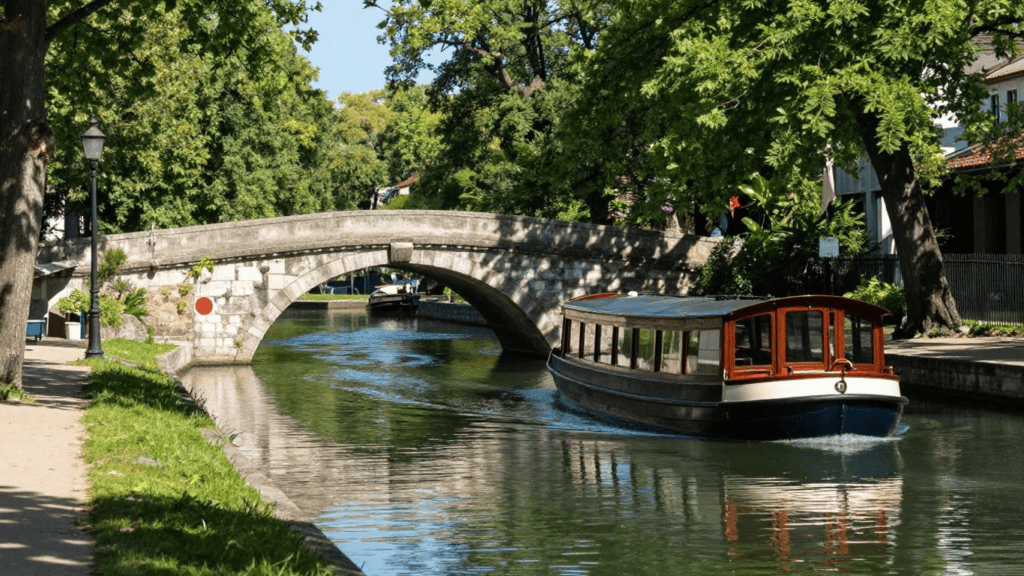 Stone arched bridge over a canal with a wooden passenger boat passing beneath, surrounded by lush green trees