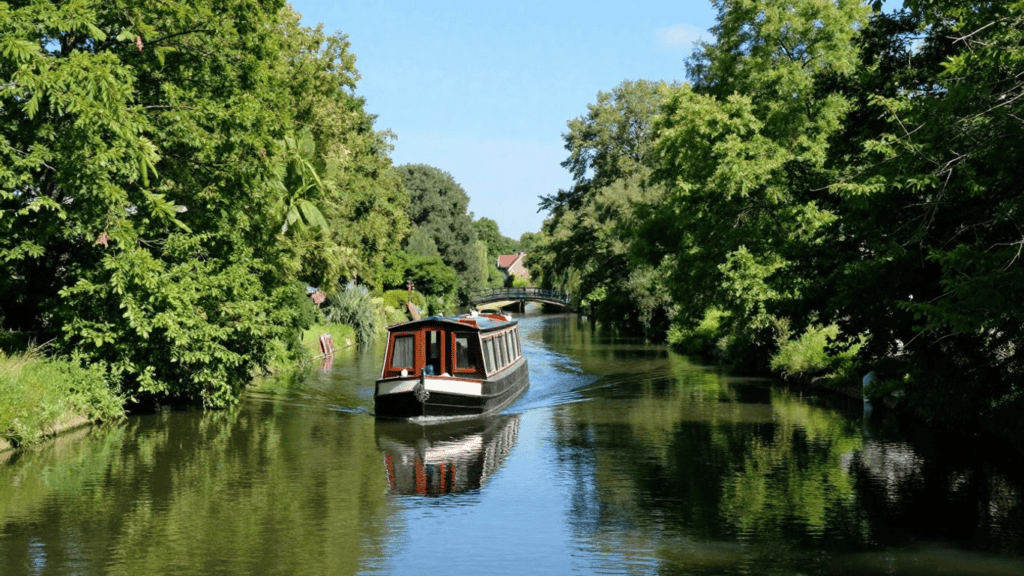 Canal boat with a wooden cabin gliding along a sunlit waterway, flanked by green trees; a small arched bridge in the distance.