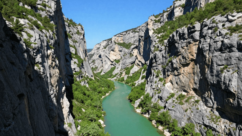 A turquoise river winds through a deep rocky canyon with steep gray cliffs and green vegetation under a clear blue sky, viewed from above
