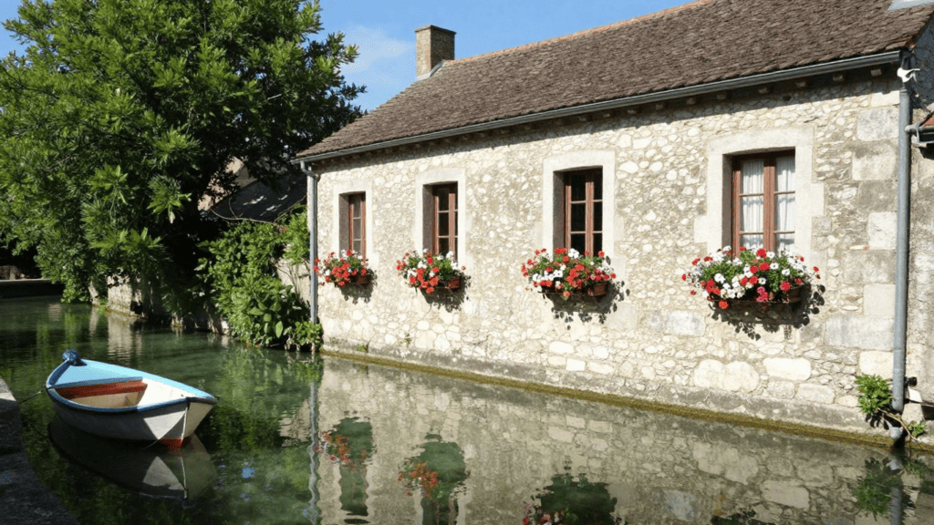 Stone cottage by a calm canal, flower-filled window boxes, and a small boat moored at the edge; reflection in the water.