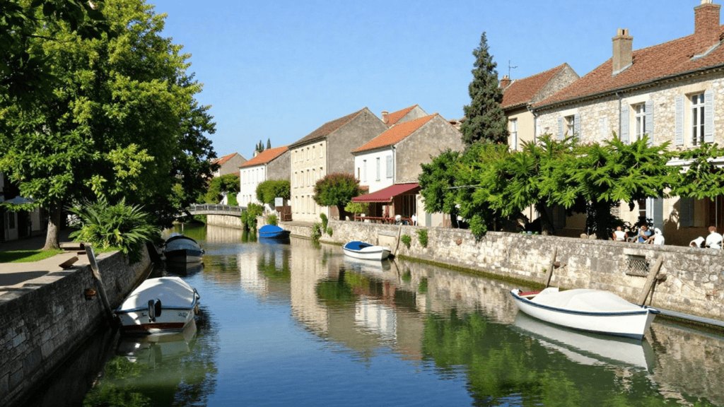 Sunny canal scene in a European village with small boats moored along stone quays and reflections in calm water across pastel stone houses with red-tiled roofs.