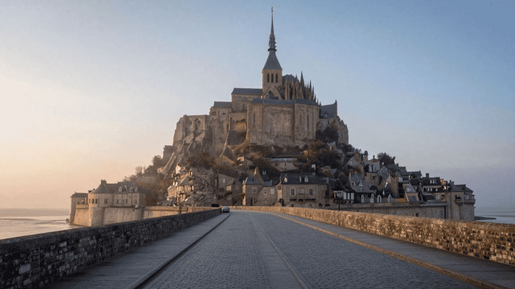 Medieval abbey and village atop Mont Saint-Michel rising from the sea, seen from the cobblestone causeway at sunset/light breeze.