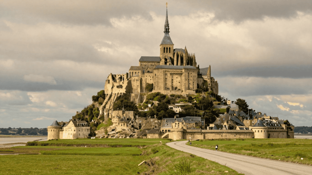 Mont Saint-Michel with its hilltop abbey and stone walls, reached by a curved causeway under a cloudy sky.
