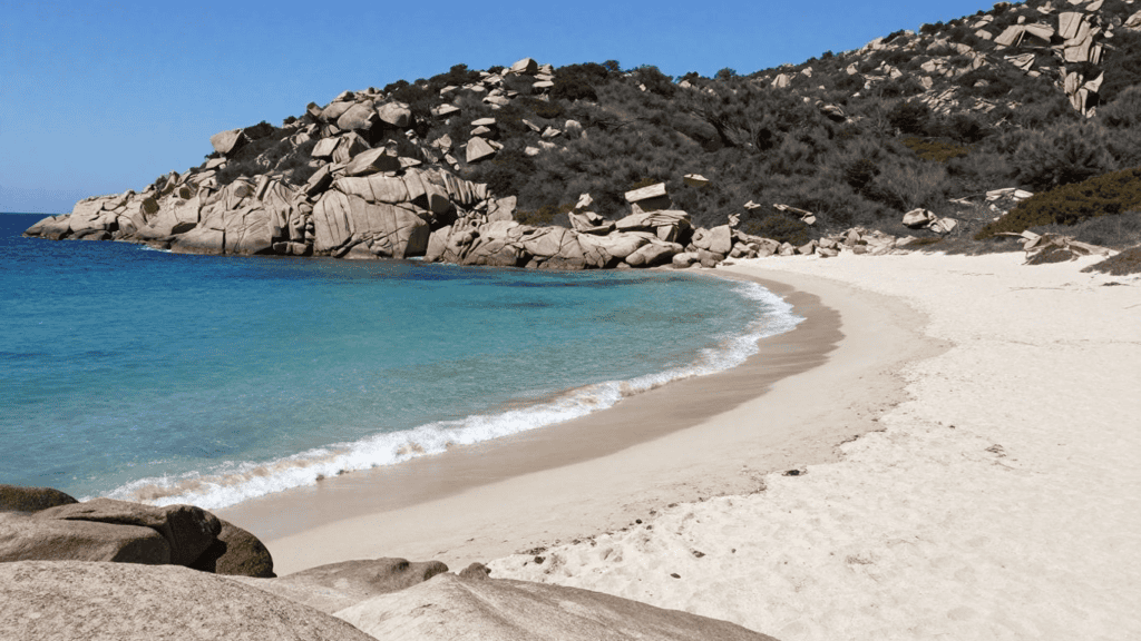 Curving white-sand beach with turquoise water and a rocky hillside under a clear blue sky.