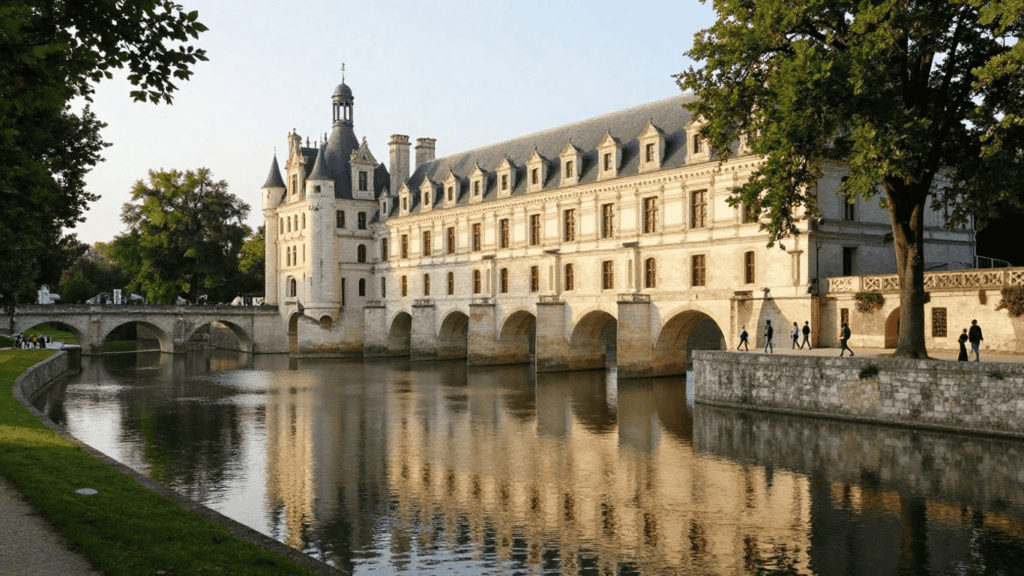 Château de Chenonceau on a stone arched bridge spanning a calm river, with trees along the banks and visitors nearby.