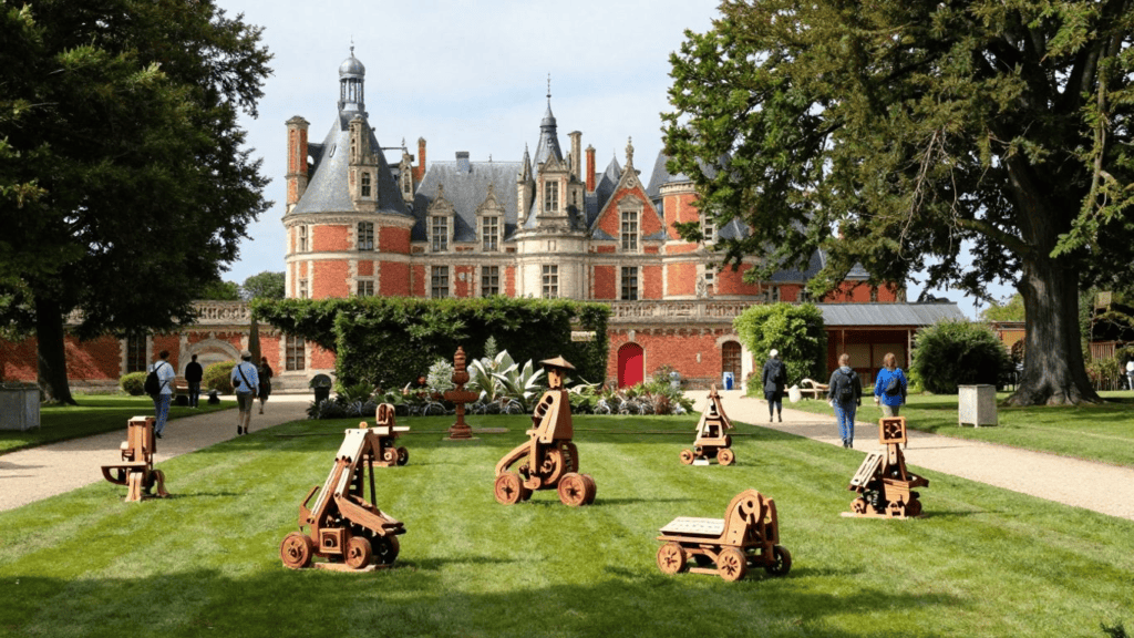 Brick château with turrets and slate roofs sits in a formal garden, wooden sculpted figures on the green lawn in the foreground.