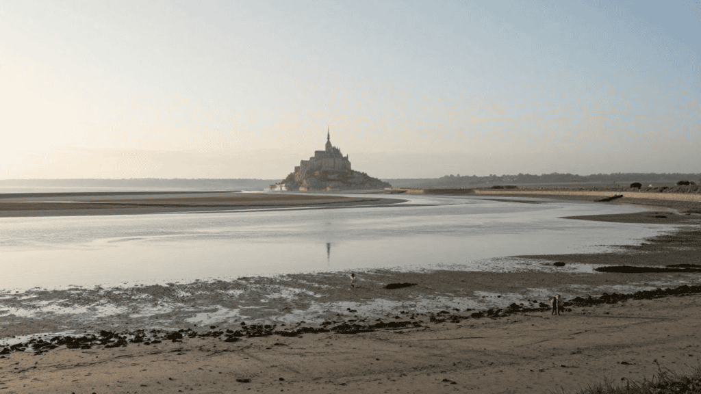 A distant view of Mont Saint-Michel rising from an tidal flat with calm water reflecting the sky and a few people on the shore nearby.