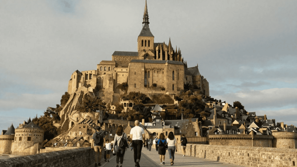 Group of visitors walking on a stone bridge toward Mont Saint-Michel’s medieval fortress on a hilltop town.
