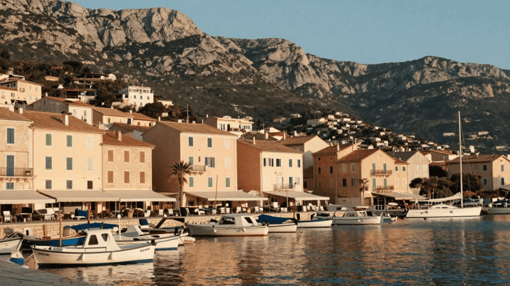 Harbor view of a Mediterranean town with pastel buildings, boats, and mountains in the background.