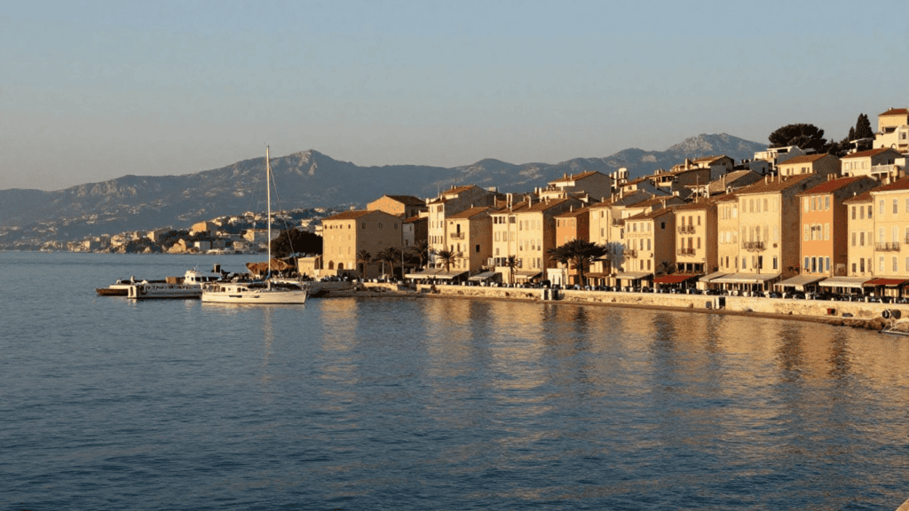 Coastal town along a calm harbor with pastel buildings, moored boats, and distant mountains.