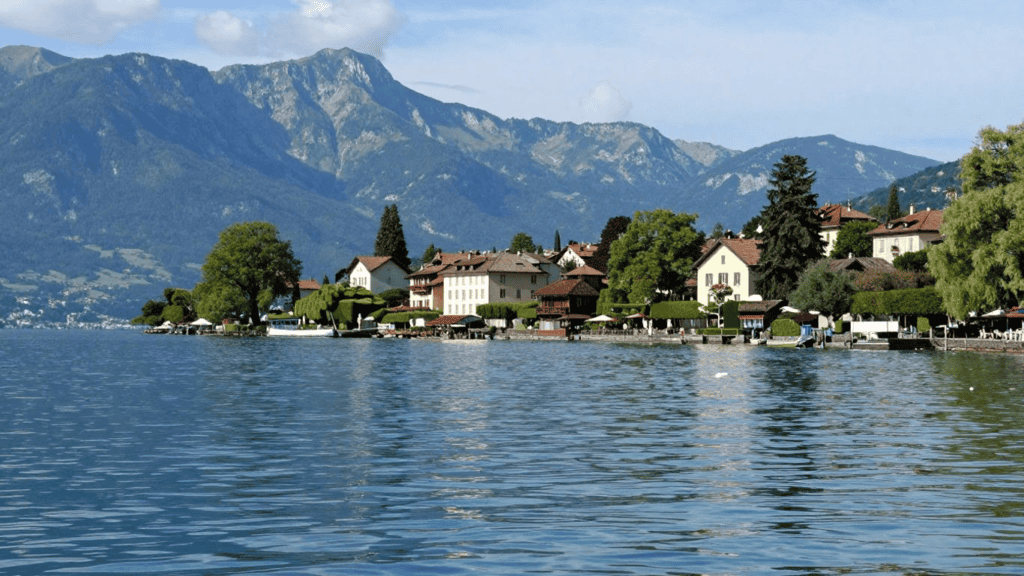 Lakeside village with pastel houses and trees along the shore, mountains rising in the distance behind. A calm lake mirrors the scene.