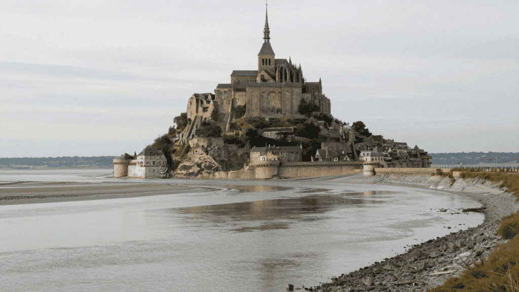 Medieval abbey and stone village on a rocky island surrounded by tidal flats and a low seawall, connected to the coast by a causeway at low tide.