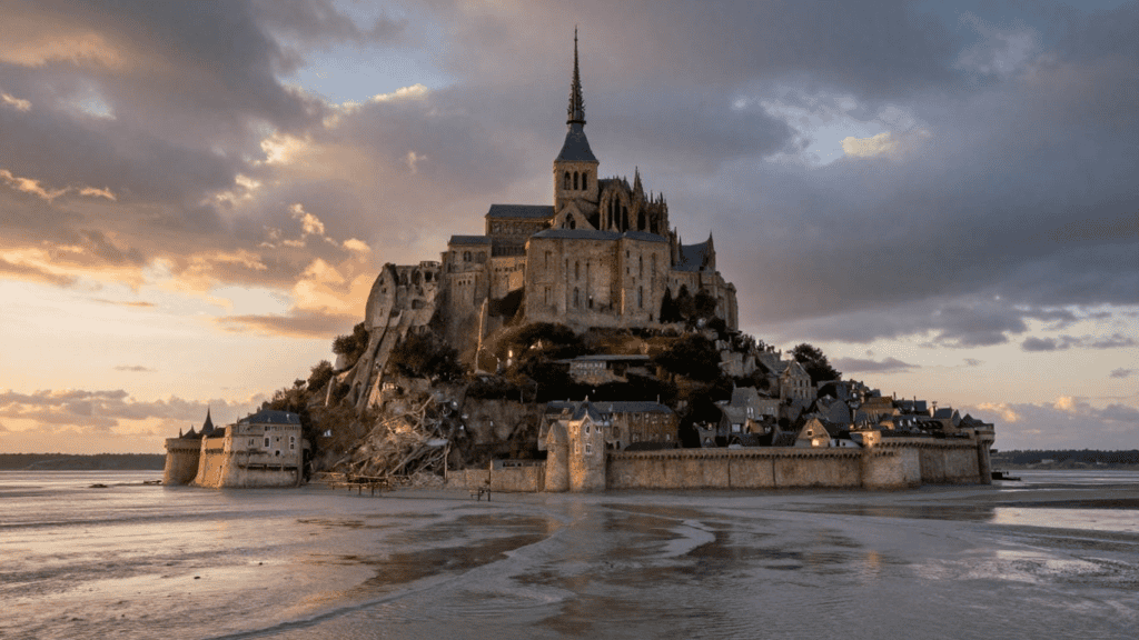 Medieval abbey atop a rocky island at Mont-Saint-Michel during sunset, with a surrounding village and tidal flats nearby.