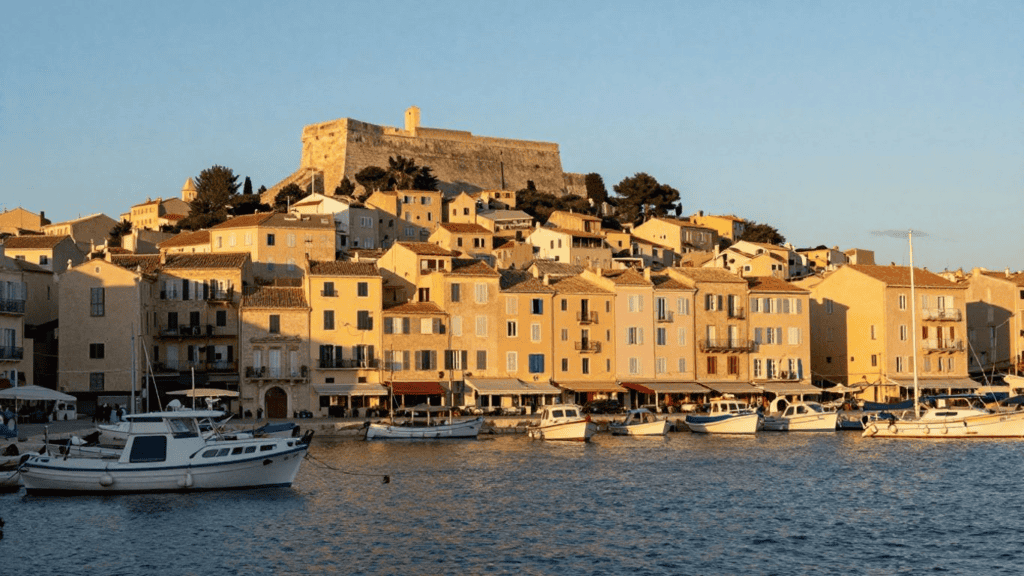 Coastal harbor town with pastel buildings lined along the water and a stone fortress on a hilltop. (Option: concise summary)