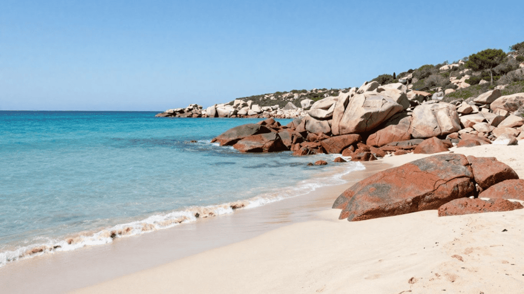 Clear blue ocean beside a sandy beach with large reddish rocks and a rocky coastline in the background.