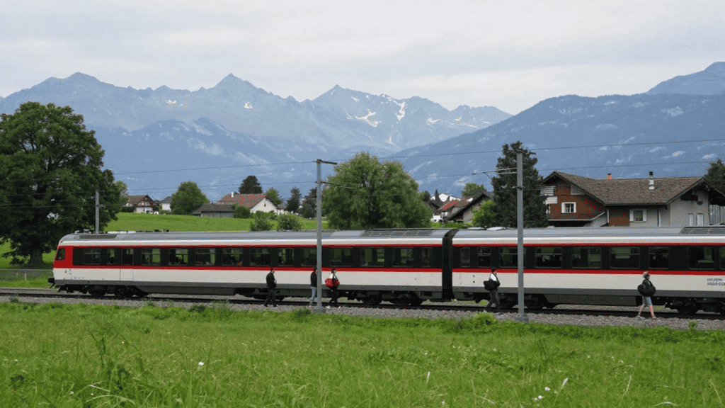 Passenger train with a red stripe passing through a green rural landscape and mountains in the distance, with people walking along the track-side