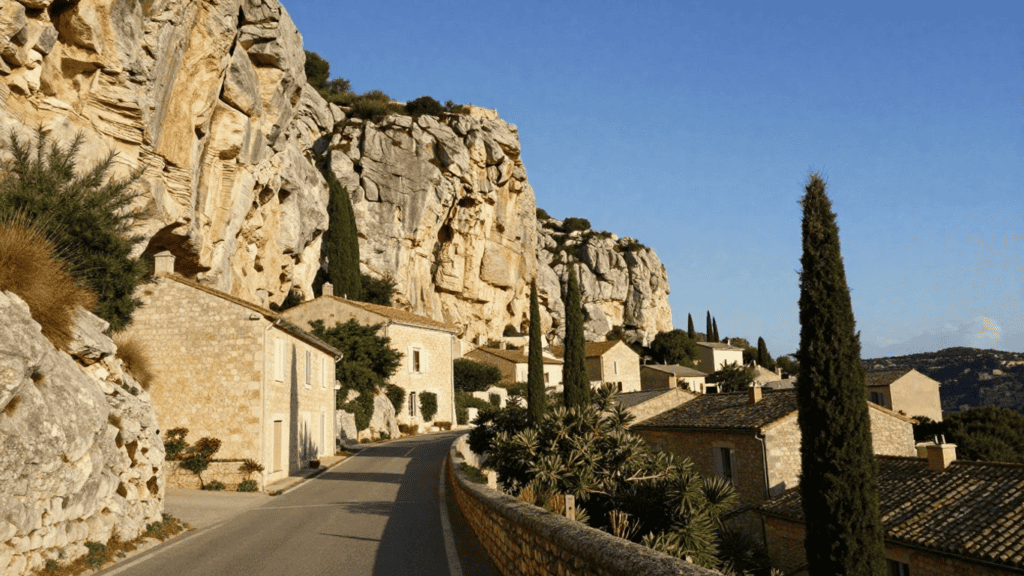 Stone houses line a winding road along a limestone cliff, with tall cypress trees under a clear blue sky.