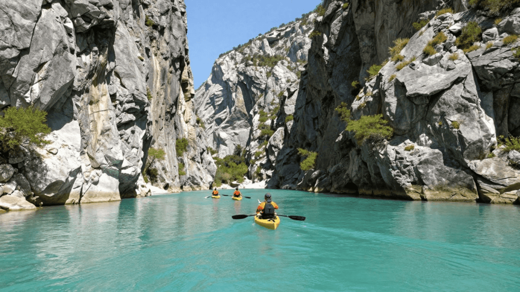 Kayakers paddle through a turquoise river between towering gray canyon walls under a clear blue sky, sunlight gleaming on the water.