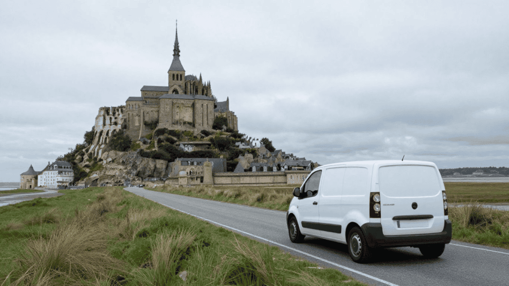 White cargo van on a coastal road approaching Mont Saint-Michel, a medieval abbey perched on a rocky island in the distance.