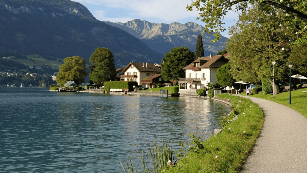 Lakeside village with houses along a calm lake, a gravel path bordered by grass, and mountains in the distance.